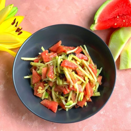 Raw Watermelon Rind and Avocado Salad seen in in the pic in a black bowl. Few pieces of watermelon slices are seen in the background and a yellow lily flower for presentation.
