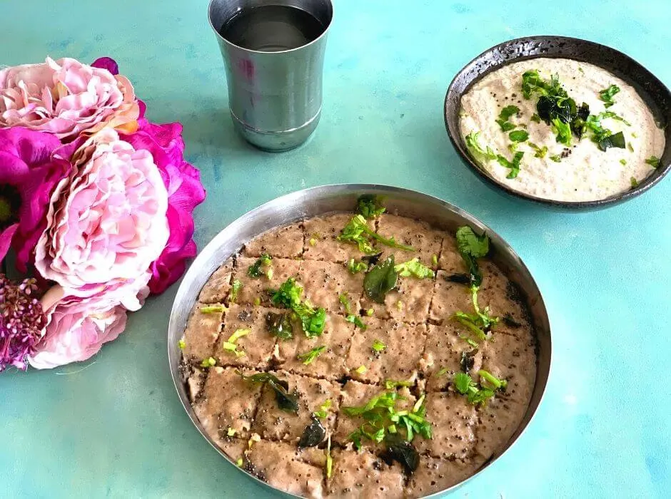 Steamed Farali Kuttu aur Singhare ke atte ka dhokla seen in a steel plate, with the tempering of mustard seeds, corainder and curry leaves, with peanut chutney as a side. Water and some flowers are seen on the side.