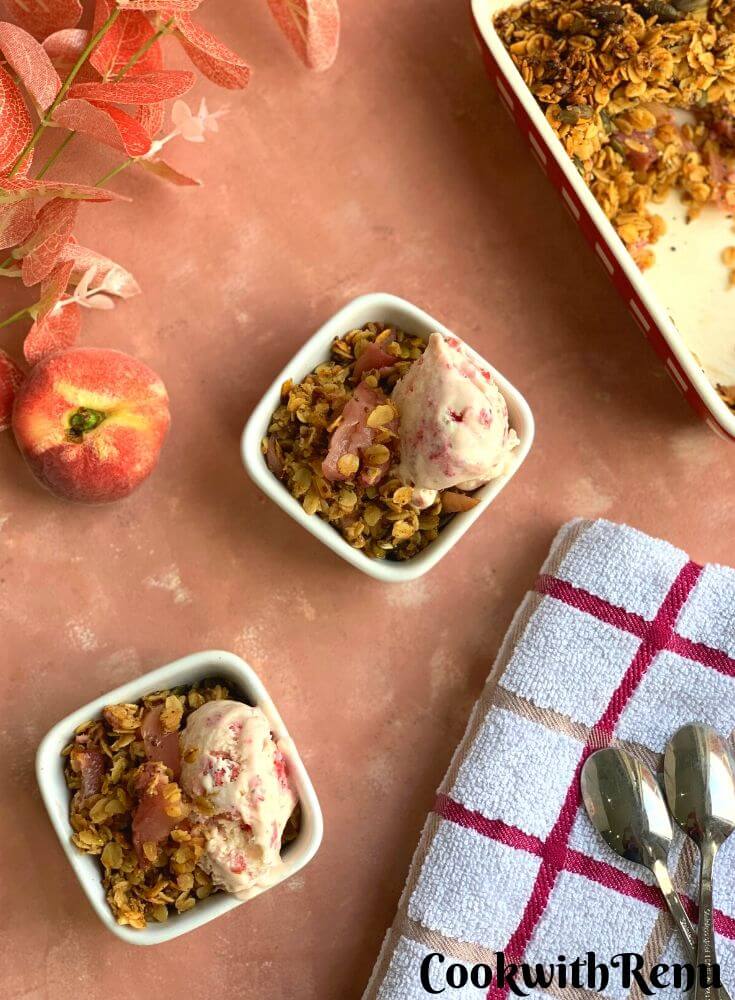 Peach Crisp served in 2 square white bowls, with Ice cream topped on it. Seen in the background is a donut peach and some flowers and a kitchen napkin on the side