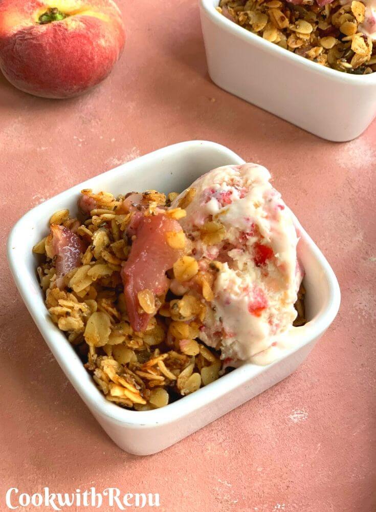 Close up look of the peach crisp served in a white rectangular bowl with icecream on top. Seen in the background is a donut peach