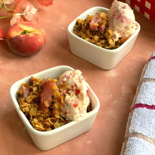 Gluten Free Peach Crisp served in 2 square white bowls, with Ice cream topped on it. Seen in the background is a donut peach and some flowers and a kitchen napkin on the side