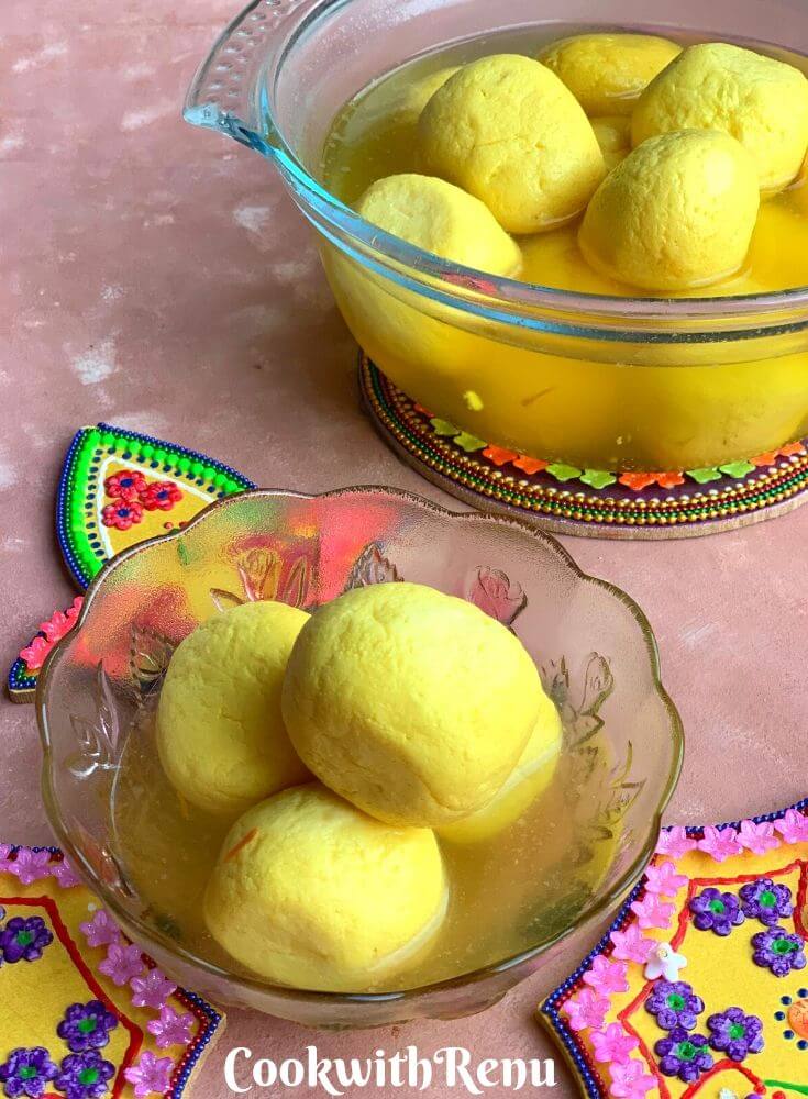 Kesar Rasgullas presented in a small glass bowl seen in the near. At the back there is a large bowl with many rasgullas.