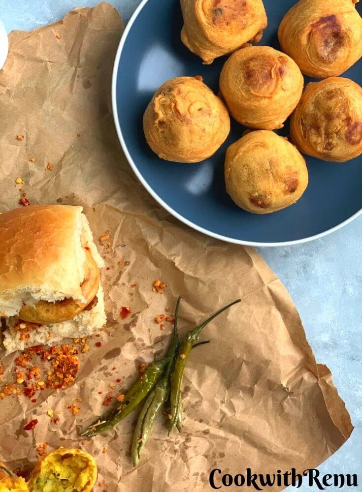 Single Vada pav being served on a paper, with red garlic chutney. Fried Green chilies are also served along and an open vada is seen in the background