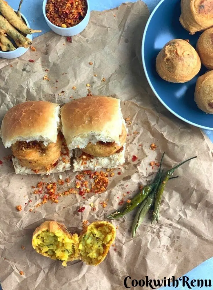 Mumbai Special Vada pav being served on a paper, with red garlic chutney. Fried Green chilies are also served along and an open vada is seen in the background
