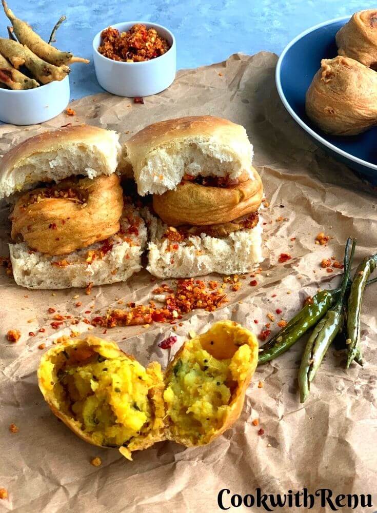 Vada pav being served on a paper, with red garlic chutney. Fried Green chilies are also served along and an open vada is seen in the background