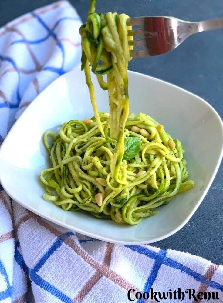 Zoodles served in a white plate. A fork with some zoodles entangled in it ready to eat. A white and blue kitchen towel seen in the background