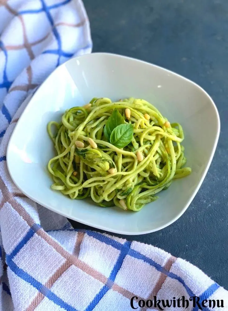 Zoodles served in a white plate, with some basil and pine nuts garnished. A white and blue kitchen towel seen in the background