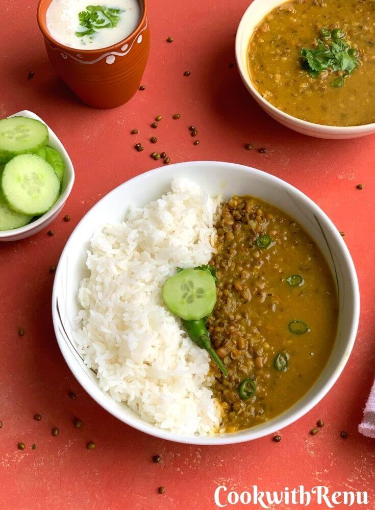 Green mung/Moong dal served in a bowl with steamed rice. A garnish of green chilly and cucumber on top. Seen along is a glass of buttermilk/chaas and another bowl of dal