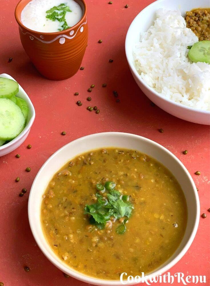Green mung/Moong dal served in a bowl with garnish of green chilly and corinader. Seen on the side are cucumber and a glass of buttermilk/chaas. A bowl of steamed rice is also seen