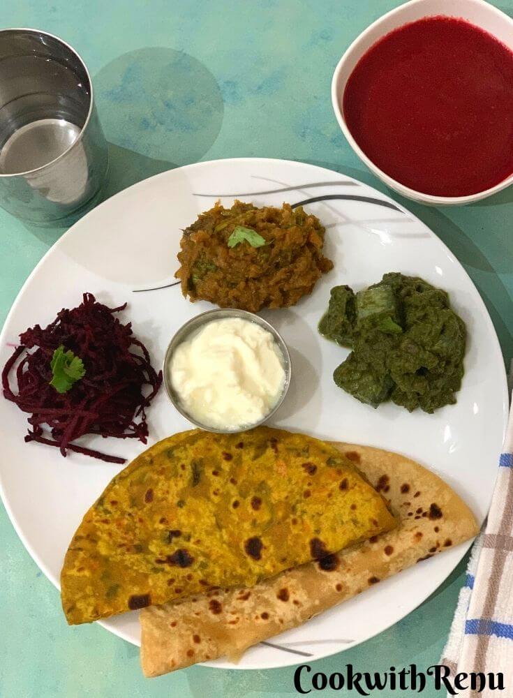 Thali has Clockwise from 6'o clock, 2 Parathas (carrot & Plain), Beetroot Salad, Turnip Bharta, Kale, tofu and corn curry and yogurt in between. Beetroot soup is seen on the side along with a glass of water on the other side.