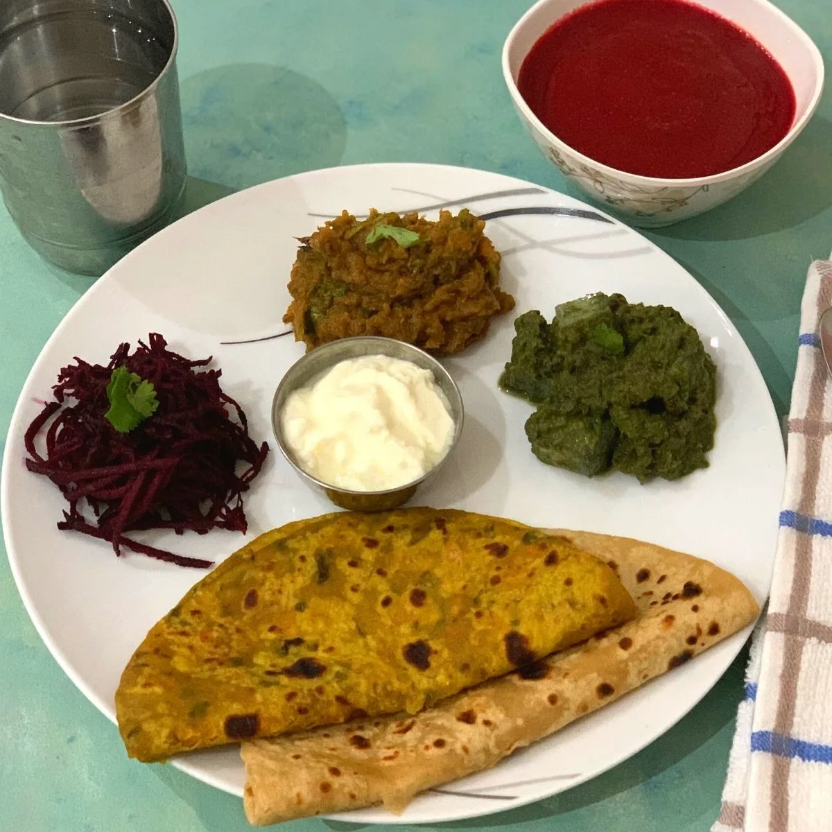 Winter Special Vegetarian Thali has Clockwise from 6'o clock, 2 Parathas (carrot & Plain), Beetroot Salad, Turnip Bharta, Kale, tofu and corn curry and yogurt in between. Beetroot soup is seen on the side along with a glass of water on the other side.