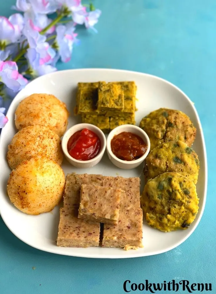 Easy Steamed Meal Platter has Four Steamed Dishes Idli, Kothimbir Vadi, Chana Dal Bafuri, and Dhokla are presented in a white square plate with Tomato chutney and tomato sauce. Seen in the background are some flowers.
