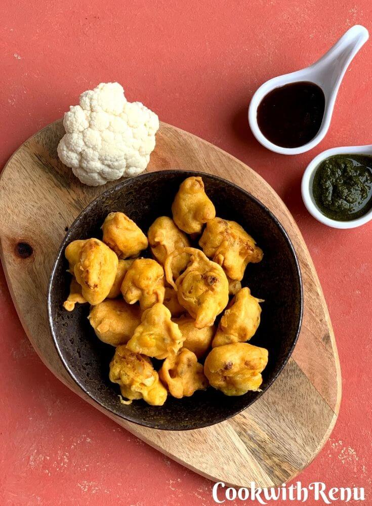 Gobi pakora or Cauliflower pakoda is an irresistible and addictive tea time fritter which are crunchy on the outside, soft inside and perfect as a party appetizer. They are served in a black bowl with 2 chutneys, coriander and tamarind.