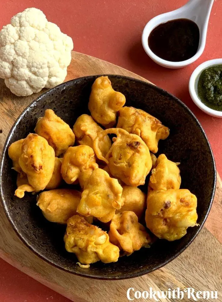 Gobi pakora or Cauliflower pakoda is an irresistible and addictive tea time fritter which are crunchy on the outside, soft inside and perfect as a party appetizer. They are served in a black bowl with 2 chutneys, coriander and tamarind.