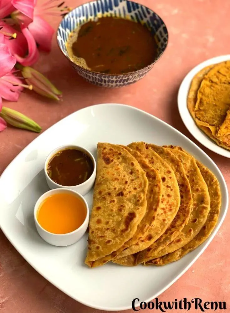 Three Puran Poli arranged on a white plate, served with ghee (Clarified butter) and katachi amti. A big bowl of amti is seen in the background and 1 puran poli which is open and the stuffin inside is seen