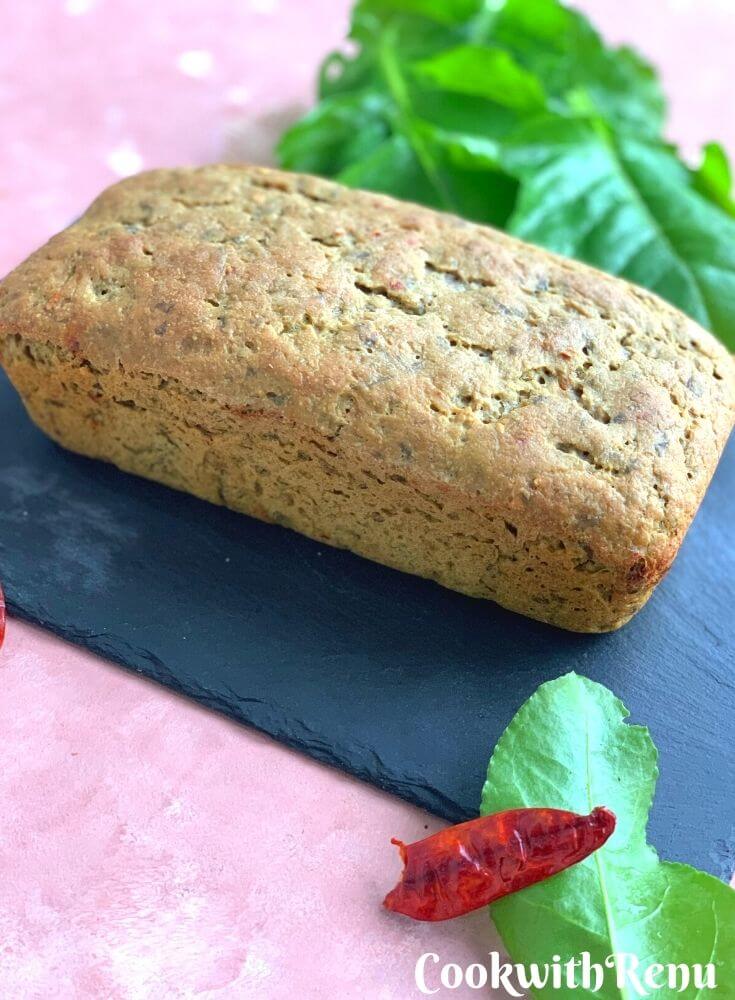 Loaf of Spinach Spiced Sourdough Discard Bread presented on a black board, with spinach and red chilly seen in background