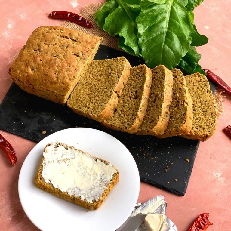 Close up look of the bread with slices cut. Texture of the bread can be seen closely with some spinach and red chilly flakes on the side. A slice of Bread with cheese spread on it is also seen
