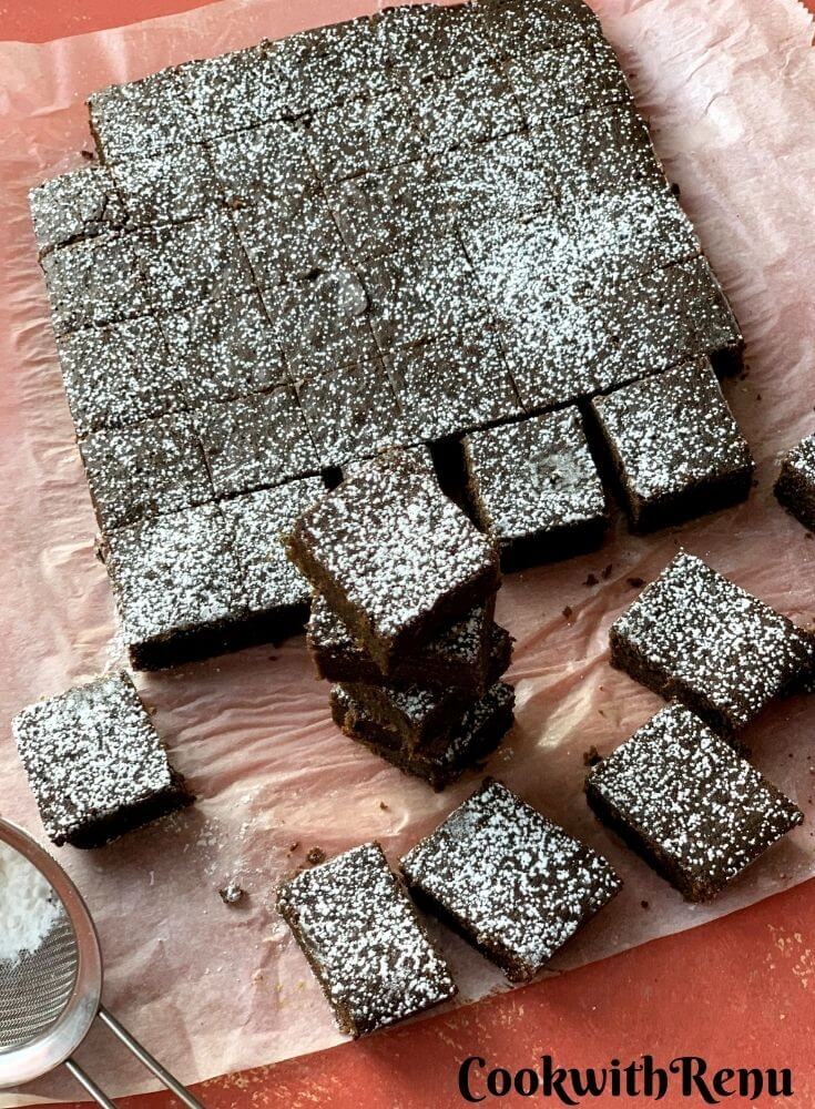 Baked brownie squares on a parchment paper, with a drizzle of icing sugar. Few brownie square are stacked one above the other.