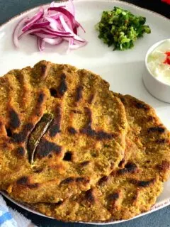 Close up look of Thalipeeth served in a white plate with yogurt, green chilly and onion.