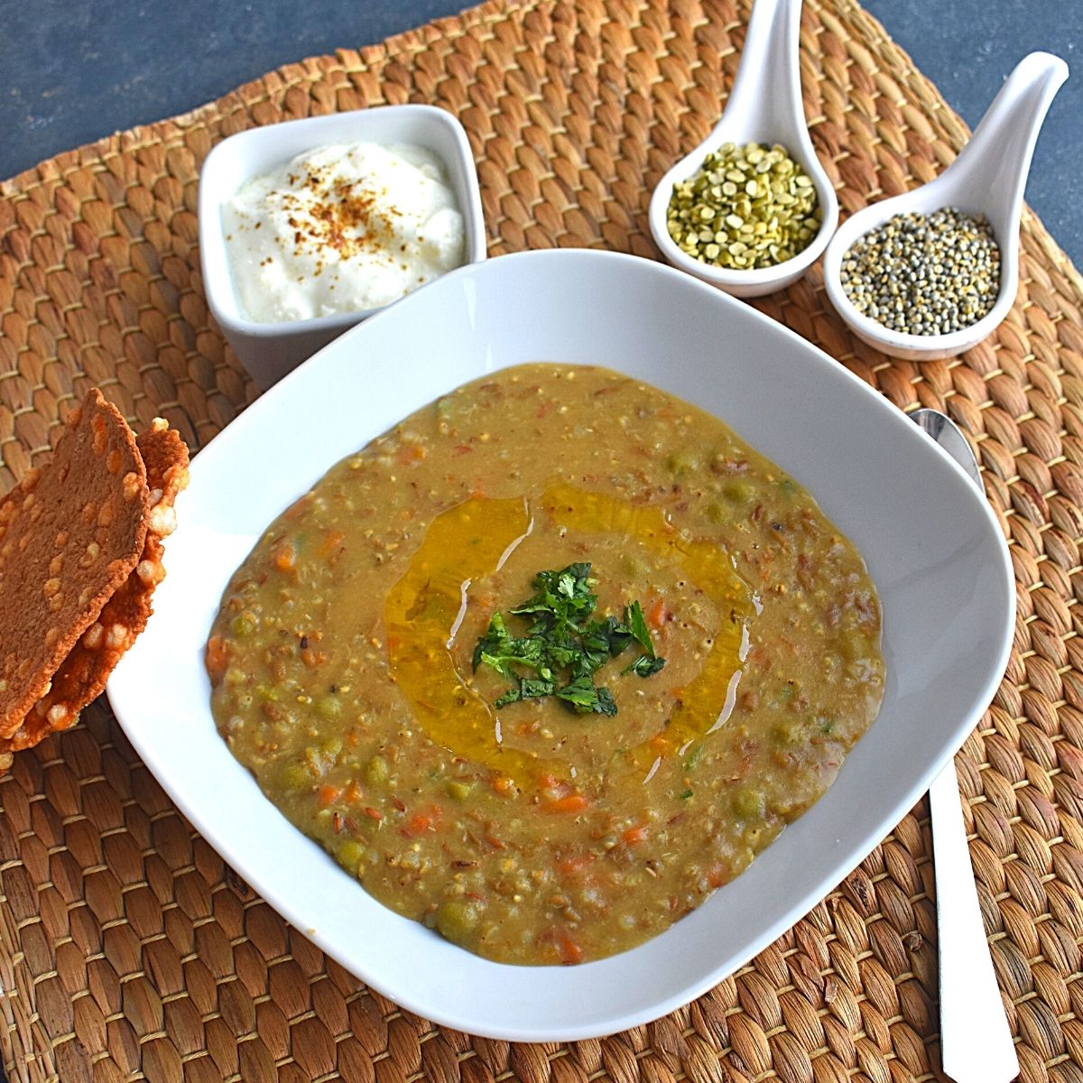Close up view of Bajra and Green Dal Khichdi served in a white rectangular bowl, with ghee drizzled on top and some coriander sprinkled. Seen in background is dal and bajra, along with dahi and sabudana (Sago seeds) papad