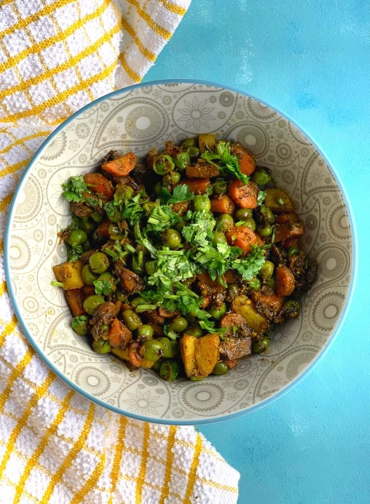 A grey bowl with Gajar Aloo Matar sabji and coriander garnished on top. Seen on the side is a yellow kitchen towel