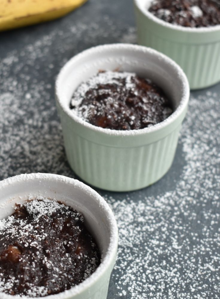 3 Chocolate Instant Pot Mug cakes seen in a line, with a banana on the side. It has a drizzle of white icing sugar