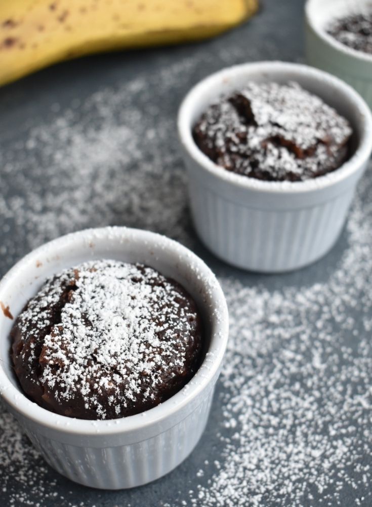 3 Chocolate Instant Pot Mug cakes seen in a line, with a banana on the side. It has a drizzle of white icing sugar