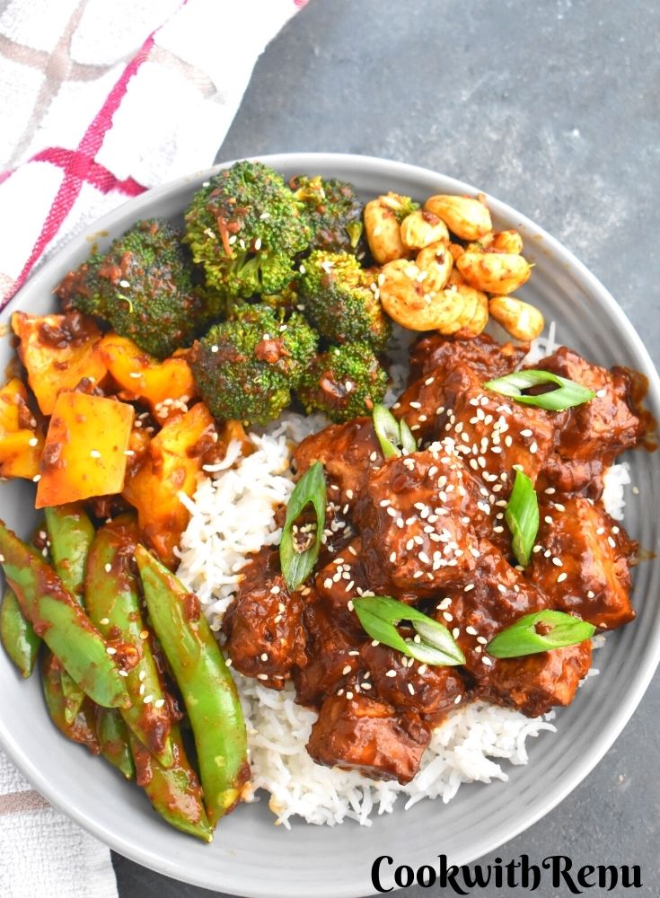 Pan-Fried Crispy Tofu served along with rice, green peas, yellow peppers, broccoli and cashew in a grey plate