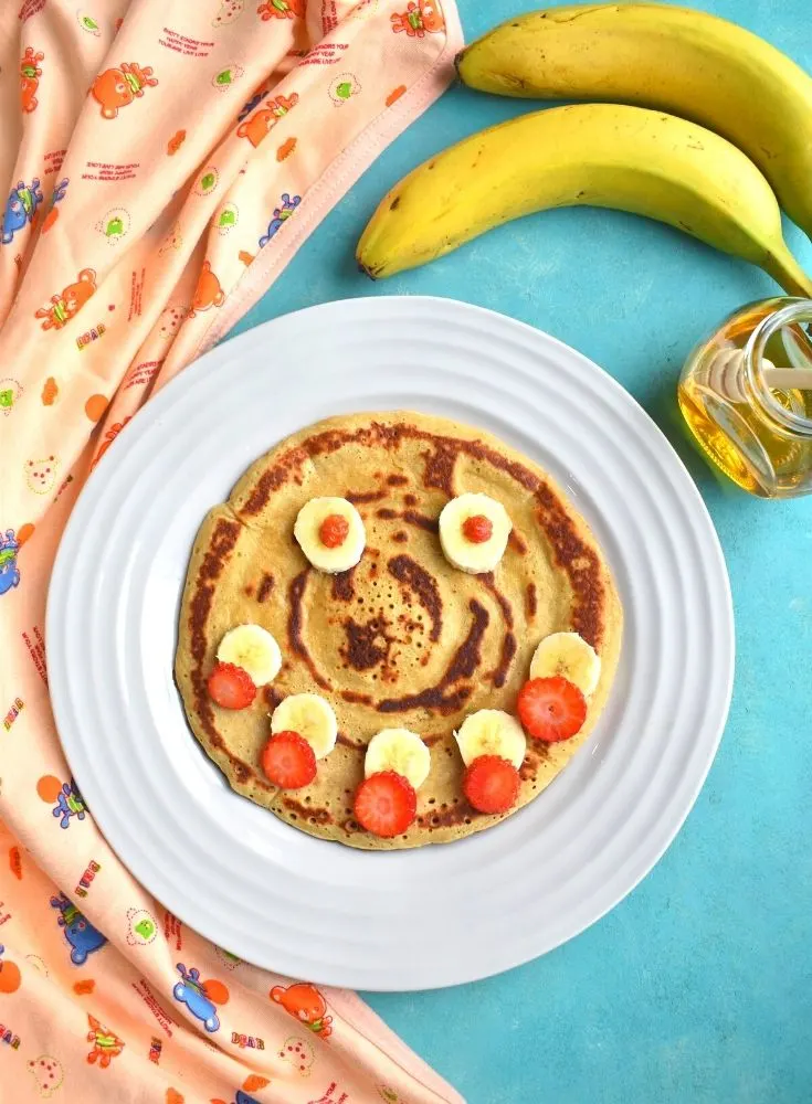 Eggless Whole Wheat Banana Pancakes with a smiley face decorated using banana and strawberries with banana and honey on the side