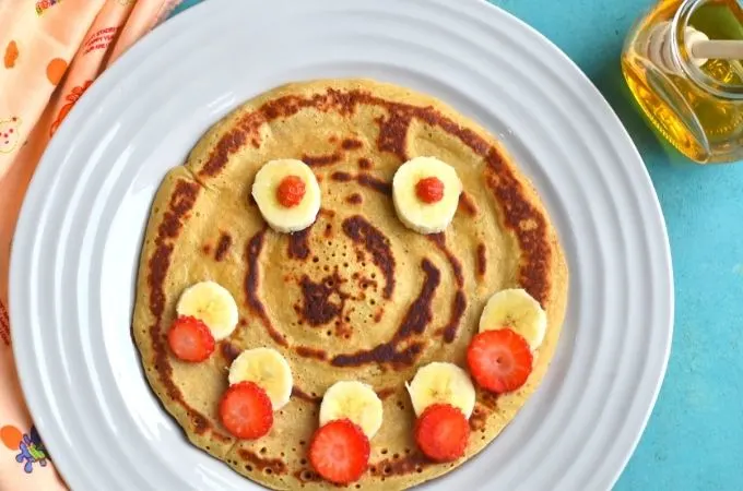 Close up look of Eggless Banana pancakes with a smiley face decorated using banana and strawberries with honey bottle on the side