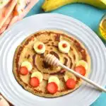 Pancake with a smiley face decorated using banana and strawberries with banana and honey on the side.