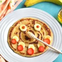 Pancake with a smiley face decorated using banana and strawberries with banana and honey on the side.