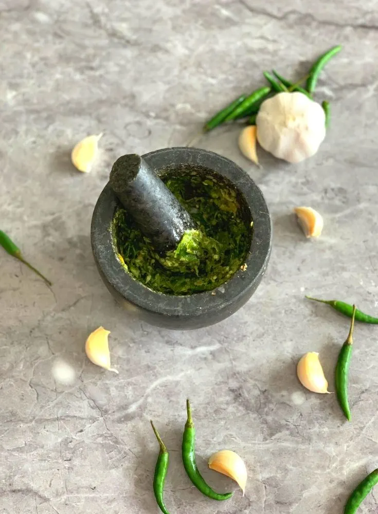 Chutney being grounded in a mortar and a pestle. Seen in the background are garlic and green chilli scattered in the background