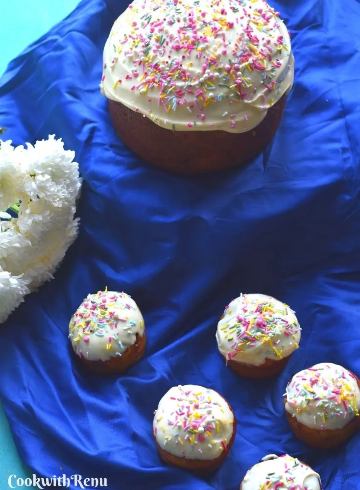 Easter Bread in round and Muffin shape, on a blue cloth with white icing and sprinkler decorations. Seen are some white flowers in the background