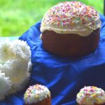 Easter Bread in round and Muffin shape, on a blue cloth with white icing and sprinkler decorations. Seen are some white flowers in the background