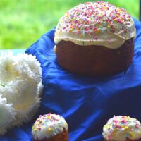 Easter Bread in round and Muffin shape, on a blue cloth with white icing and sprinkler decorations. Seen are some white flowers in the background