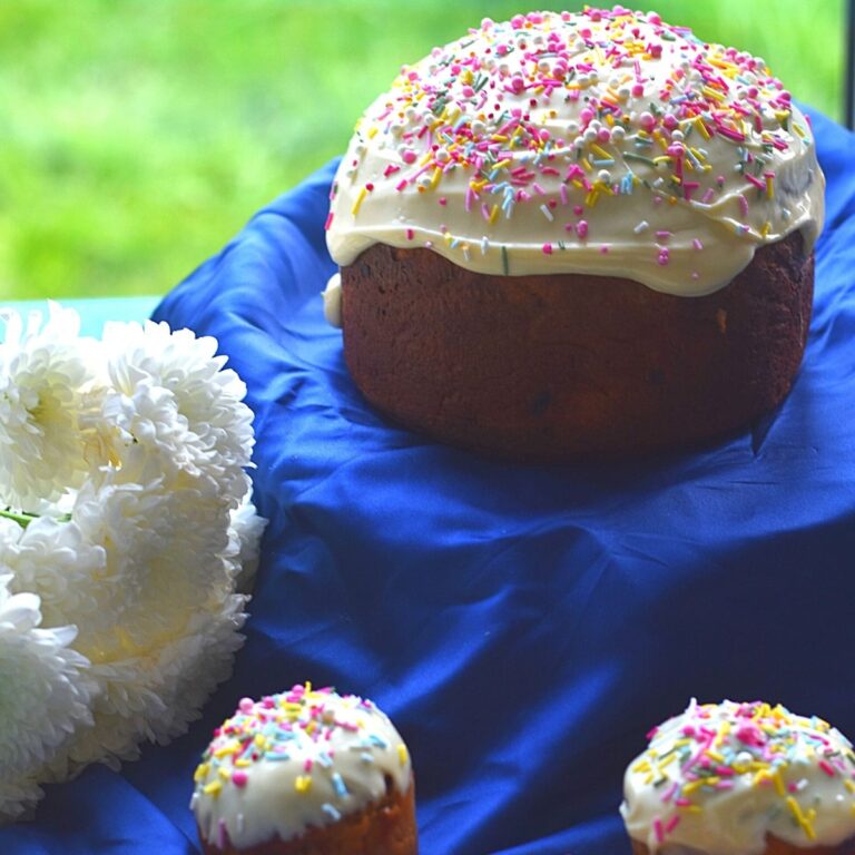 Easter Bread in round and Muffin shape, on a blue cloth with white icing and sprinkler decorations. Seen are some white flowers in the background