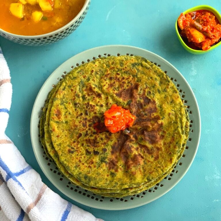 Kale Paratha stacked up on a plate with a dollop of pickle on the top. Seen in the background is some potato tomato curry and pickle