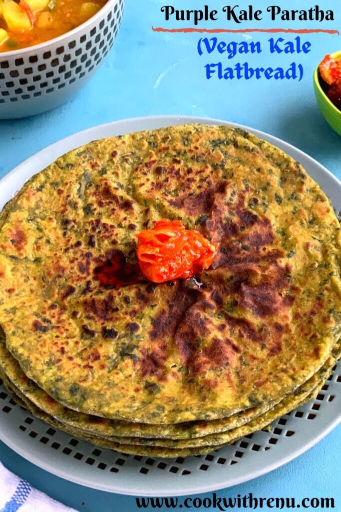 Kale Paratha stacked up on a plate with a dollop of pickle on the top. Seen in the background is some potato tomato curry and pickle