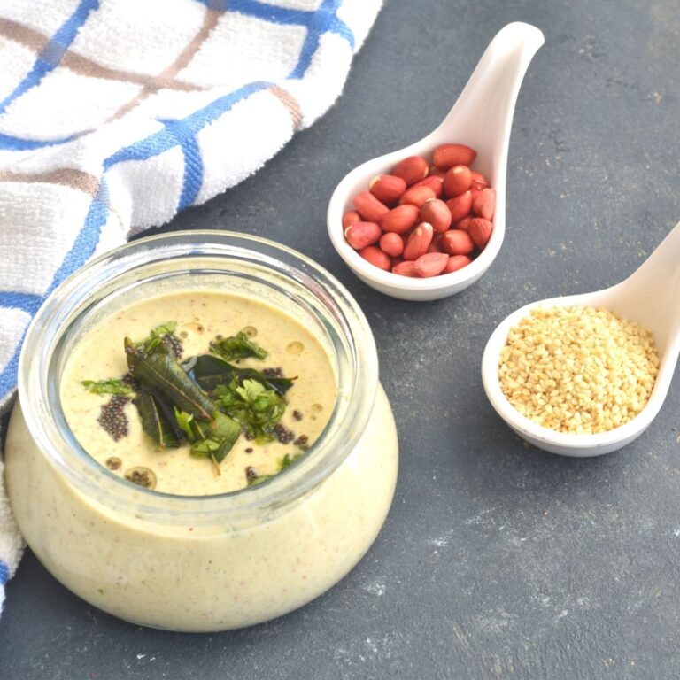Farali Chutney in a glass jar with a tempering of mustard seeds and curry leaves. Seen in the background is a small bowl or peanuts and sesame seeds and a blue and white napkin