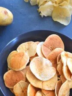 Fried Sun-Dried Potato Chips served in a blue bowl, with uncooked potato chips and 2 potatoes seen in background