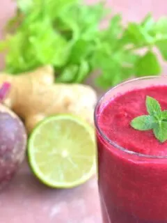 Ginger, Beetroot & Celery Juice served in 2 glasses with a garnish of mint. Seen in the background is some celery leaves, beetroot, ginger and lemon