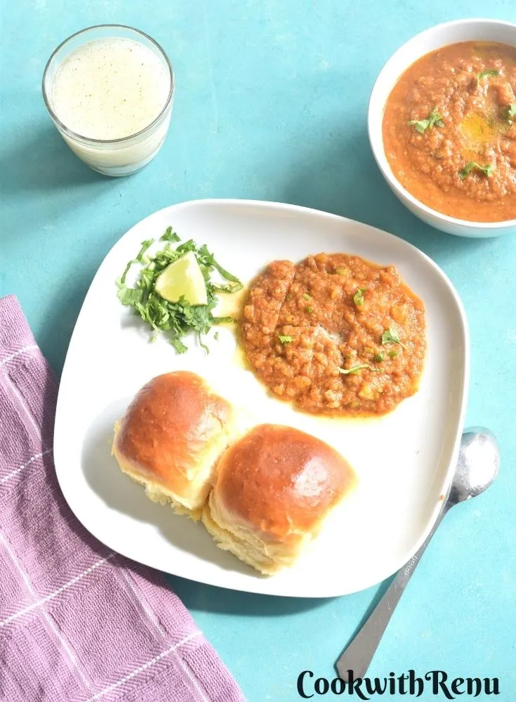 Pav Bhaji served on a white plate with 2 pav, coriander and lemon. Seen in the background is a glass of buttermilk and a bowl full of pav bhaji
