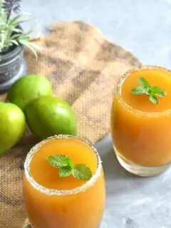 Aam Panna with Jaggery served in 2 glasses with a mint garnish. Seen along side are some green mangoes