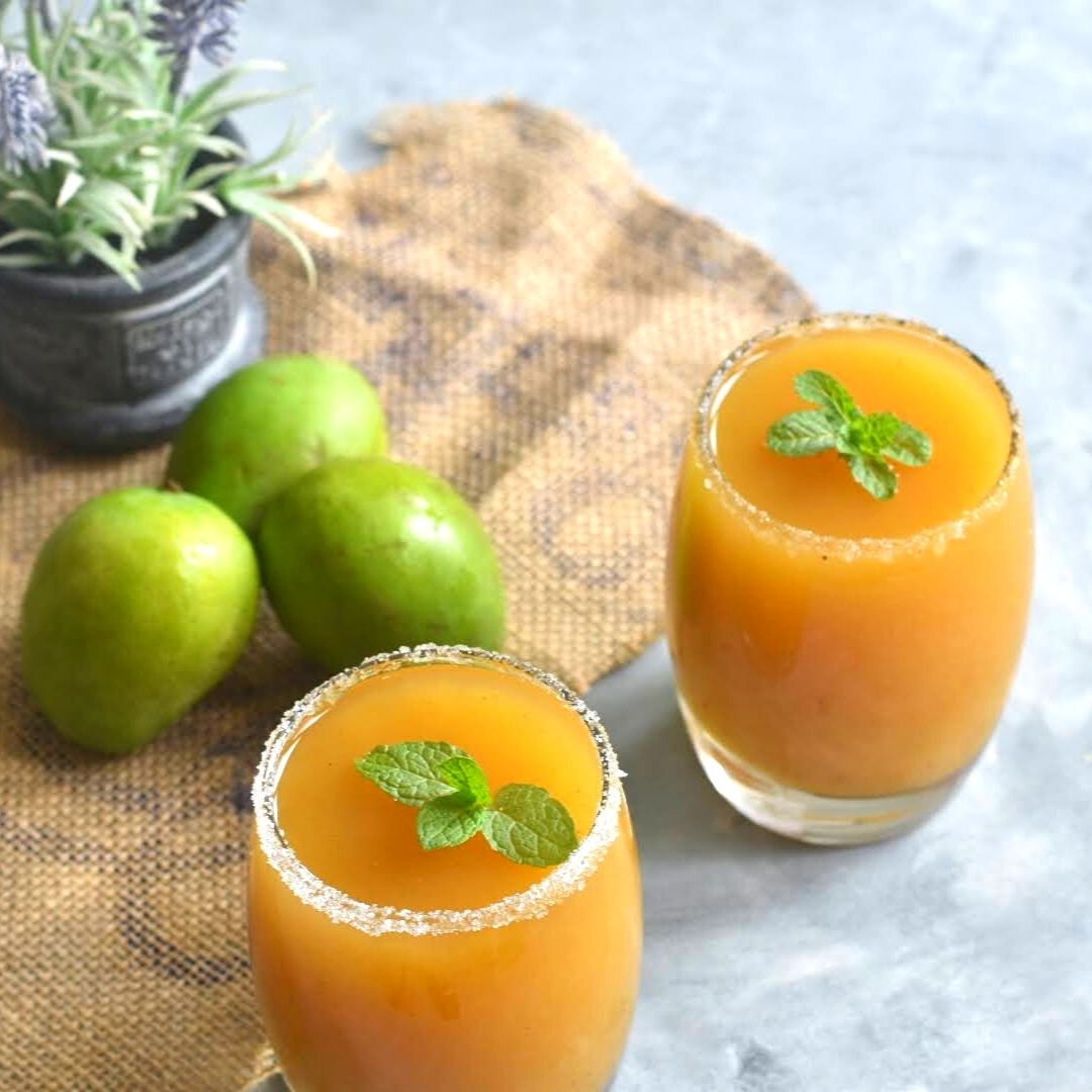 Aam Panna with Jaggery served in 2 glasses with a mint garnish. Seen along side are some green mangoes