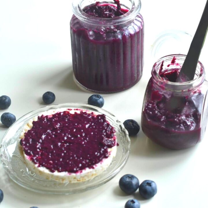 Blueberry chia jam spread on a rice cracker. Seen are 2 bottles of jar filled with jam along with some berries spread across.