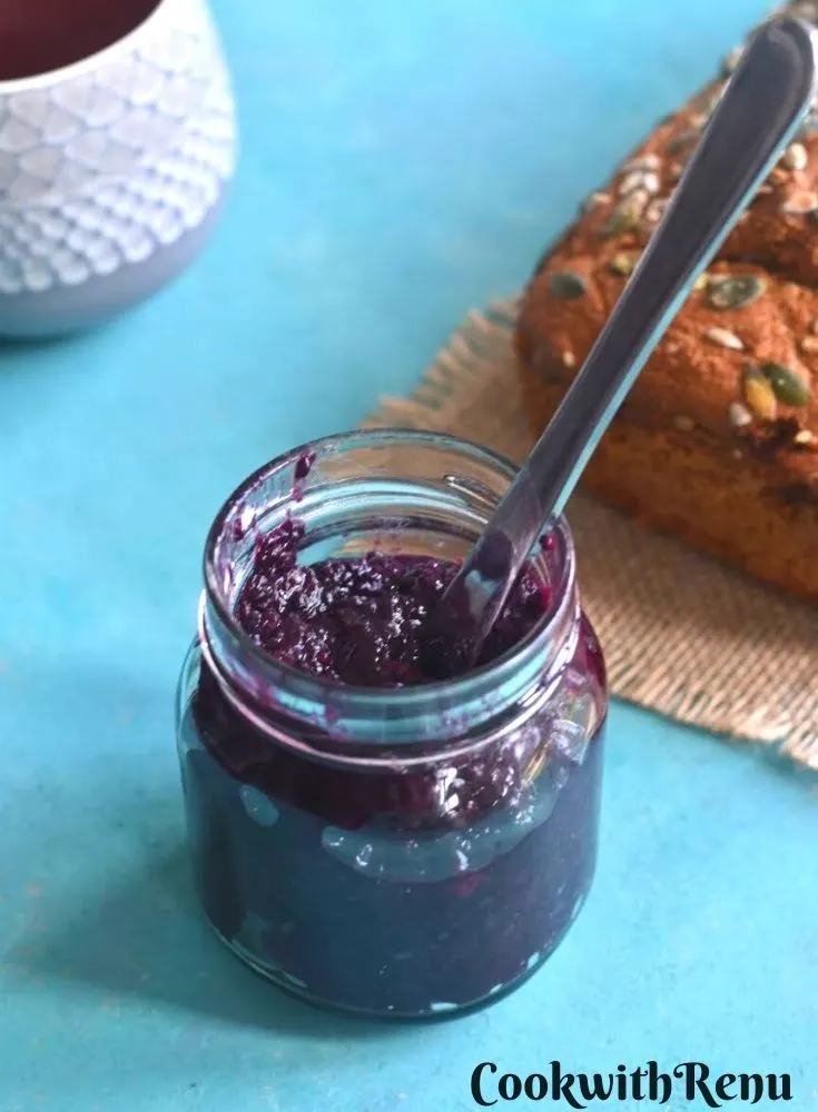 Jam filled in a glass bottle. Seen in the background is a loaf of bread a coffee mug.