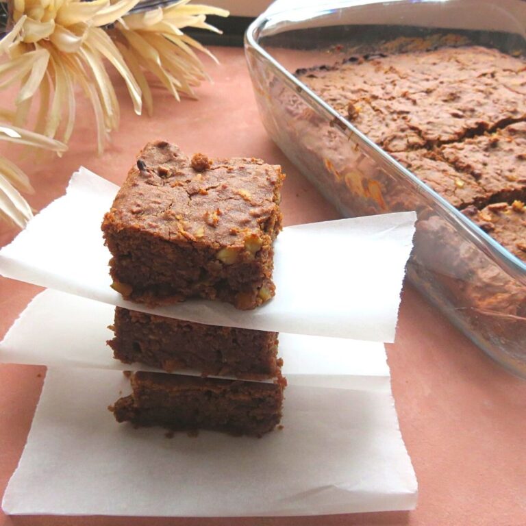 Apple brownies stacked one above the other. Seen along side is a tray of baked apple brownie along with some flowers.