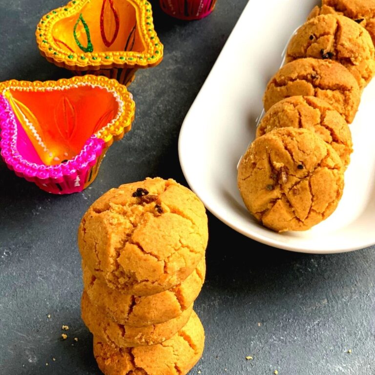 A stack of nankhatai seen , few on a white tray and seen alongside are colourful Diwali diyas