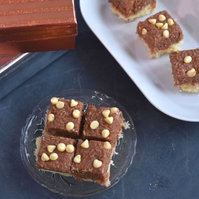 4 pieces of Chocolate Coconut Ice served on a small glass plate. Seen along are some festive gift boxes and a few more slices of barfi on a white tray